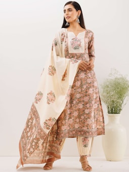 A woman stands wearing a traditional floral embroidered outfit consisting of a kurta, pants, and a dupatta. The ensemble features intricate patterns and soft, pastel colors. She accessorizes with large earrings and sandals, standing beside a white decorative vase with green foliage.
