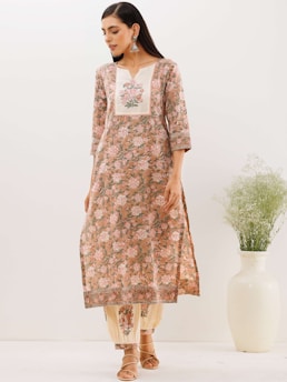 A woman is wearing a floral-patterned traditional dress with a light pink and beige color scheme. She has long dark hair and is accessorizing with large earrings and sandals. There is a large white vase with green foliage beside her.