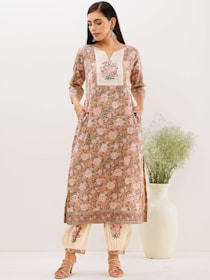 A woman is dressed in a floral-patterned traditional outfit with a long tunic and matching pants. The outfit features intricate floral designs in shades of pink and cream. Her hands are in the pockets of the tunic, and she is wearing heeled sandals. A large white ceramic vase filled with greenery stands beside her.