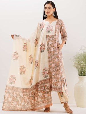 A woman is wearing a traditional floral printed outfit, consisting of a long kurta, pants, and a dupatta. The dupatta is light-colored with a floral pattern that matches the kurta. She is standing with one hand on her hip and the other holding out a part of the dupatta. The background is off-white and there is a potted plant to her side.
