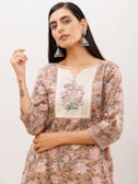 A person wearing a floral-patterned dress with intricate embroidery and large silver earrings. The dress is adorned with pink and green floral designs, and there is a tattoo visible on one wrist. The person's hand is raised near their face in a poised manner.
