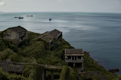Abandoned buildings covered in lush green vegetation are situated on a hillside overlooking a calm sea with a few ships visible in the distance. The structures appear weathered and overgrown, blending with the natural landscape. The scene conveys a sense of solitude and nature reclaiming human constructions.