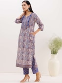 A woman stands wearing a colorful, paisley-patterned kurta with blue, purple, and white hues. She pairs it with matching printed trousers and beige heeled sandals. Her hair is long and dark, and she accessorizes with large, ornate earrings. There is a white vase with green plants on the right.