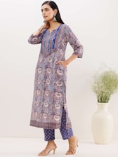 A woman stands wearing a colorful, paisley-patterned kurta with blue, purple, and white hues. She pairs it with matching printed trousers and beige heeled sandals. Her hair is long and dark, and she accessorizes with large, ornate earrings. There is a white vase with green plants on the right.