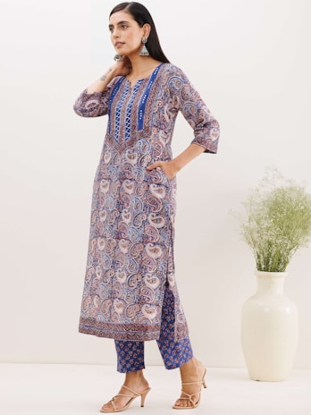 A woman stands wearing a colorful, paisley-patterned kurta with blue, purple, and white hues. She pairs it with matching printed trousers and beige heeled sandals. Her hair is long and dark, and she accessorizes with large, ornate earrings. There is a white vase with green plants on the right.