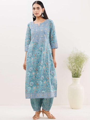 A woman is wearing a traditional blue kurta with intricate floral patterns and matching pants. She is accessorized with large earrings and stands near a white vase with greenery. The setting is minimalistic and indoors.