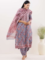 A woman is wearing a beautifully patterned traditional outfit with floral and abstract designs in shades of blue and red. She is standing with her hands clasped, wearing earrings, and has long hair. The outfit includes a kurta, trousers, and a dupatta draped around her shoulders. She is posing near a large white vase with green foliage.