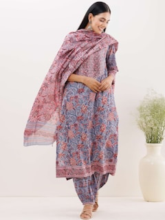 A woman is wearing a beautifully patterned traditional outfit with floral and abstract designs in shades of blue and red. She is standing with her hands clasped, wearing earrings, and has long hair. The outfit includes a kurta, trousers, and a dupatta draped around her shoulders. She is posing near a large white vase with green foliage.