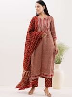 A woman wearing a traditional ethnic outfit with a long patterned red kurta and matching pants. The outfit features intricate geometric patterns and designs. She is also draped with a matching dupatta over her shoulder. The woman is standing near a large white vase containing greenery, and she is wearing hoop earrings and high heels.