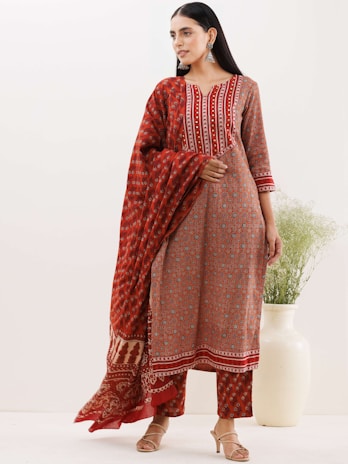 A woman wearing a traditional ethnic outfit with a long patterned red kurta and matching pants. The outfit features intricate geometric patterns and designs. She is also draped with a matching dupatta over her shoulder. The woman is standing near a large white vase containing greenery, and she is wearing hoop earrings and high heels.