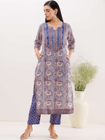 A woman is standing, wearing an ethnic printed outfit with blue, red, and white patterns. The outfit consists of a long top and pants. The woman has her hands in the pockets of the top and is wearing beige sandals. She has long, straight hair and is wearing large earrings. Behind her is a large white vase with green foliage.