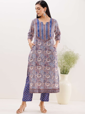 A woman is standing, wearing an ethnic printed outfit with blue, red, and white patterns. The outfit consists of a long top and pants. The woman has her hands in the pockets of the top and is wearing beige sandals. She has long, straight hair and is wearing large earrings. Behind her is a large white vase with green foliage.