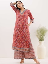 A woman is wearing an elegant red floral-patterned kurta and palazzo set with intricate designs and printed borders. She is posing with one hand on her head, showcasing her statement earrings. She stands next to a large white vase with green foliage in a minimalist indoor setting.