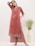 A woman is wearing an elegant red floral-patterned kurta and palazzo set with intricate designs and printed borders. She is posing with one hand on her head, showcasing her statement earrings. She stands next to a large white vase with green foliage in a minimalist indoor setting.