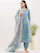 A woman is wearing a traditional blue printed outfit with intricate patterns. The ensemble includes a long top, matching pants, and a flowing dupatta. She accessorizes with large earrings and stands in a minimalistic setting with a white background and a decorative vase with green foliage.