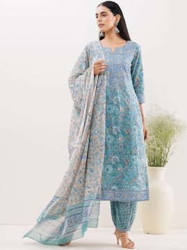 A woman is wearing a traditional blue printed outfit with intricate patterns. The ensemble includes a long top, matching pants, and a flowing dupatta. She accessorizes with large earrings and stands in a minimalistic setting with a white background and a decorative vase with green foliage.