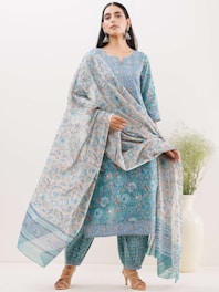 A woman stands wearing a traditional blue and beige outfit with intricate patterns. She holds a matching dupatta gracefully draped around her. The outfit features floral designs and is complemented by ethnic earrings and beige sandals. In the background, a white vase with green foliage adds a subtle decorative element.