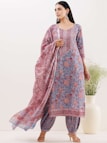 A woman is wearing a traditional outfit with intricate floral patterns in shades of blue and red. The ensemble includes a kurta, dupatta, and pants, with the dupatta elegantly draped over her shoulder. The background features a minimalistic setting with a large white vase containing green plants.