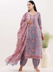 A woman is wearing a traditional outfit with intricate floral patterns in shades of blue and red. The ensemble includes a kurta, dupatta, and pants, with the dupatta elegantly draped over her shoulder. The background features a minimalistic setting with a large white vase containing green plants.