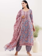 A woman is wearing a traditional outfit with intricate floral patterns in shades of blue and red. The ensemble includes a kurta, dupatta, and pants, with the dupatta elegantly draped over her shoulder. The background features a minimalistic setting with a large white vase containing green plants.