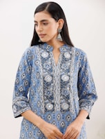 A person with long dark hair is wearing a blue patterned kurta with intricate floral designs. The attire features silver and white motifs, and the person accessorizes with large, ornate silver earrings. The background is simple and neutral, drawing focus to the clothing and jewelry.