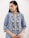 A person with long dark hair is wearing a blue patterned kurta with intricate floral designs. The attire features silver and white motifs, and the person accessorizes with large, ornate silver earrings. The background is simple and neutral, drawing focus to the clothing and jewelry.