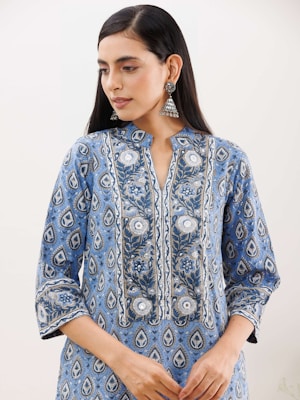 A person with long dark hair is wearing a blue patterned kurta with intricate floral designs. The attire features silver and white motifs, and the person accessorizes with large, ornate silver earrings. The background is simple and neutral, drawing focus to the clothing and jewelry.