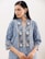 A person with long dark hair is wearing a blue patterned kurta with intricate floral designs. The attire features silver and white motifs, and the person accessorizes with large, ornate silver earrings. The background is simple and neutral, drawing focus to the clothing and jewelry.