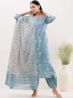 A woman is wearing a traditional blue and beige kurta with intricate floral patterns. The outfit includes a matching dupatta draped elegantly over her shoulders. She accessorizes with large, decorative earrings and wears high-heeled sandals. A large white vase with green plant stems is placed to the right.