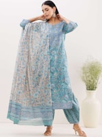 A woman is wearing a traditional blue and beige kurta with intricate floral patterns. The outfit includes a matching dupatta draped elegantly over her shoulders. She accessorizes with large, decorative earrings and wears high-heeled sandals. A large white vase with green plant stems is placed to the right.