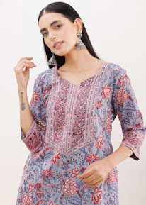 A person wearing a floral-patterned kurta with vibrant colors of red, pink, blue, and gray. The outfit features intricate embroidery along the neckline. The individual is accessorized with large, ornate silver earrings and has long, dark hair. The person has a tattoo on their forearm.