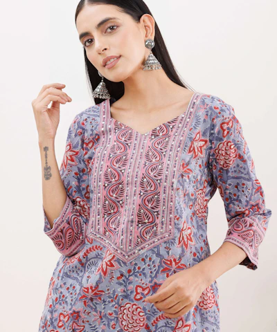 A person wearing a floral-patterned kurta with vibrant colors of red, pink, blue, and gray. The outfit features intricate embroidery along the neckline. The individual is accessorized with large, ornate silver earrings and has long, dark hair. The person has a tattoo on their forearm.