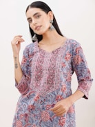 A person wearing a floral-patterned kurta with vibrant colors of red, pink, blue, and gray. The outfit features intricate embroidery along the neckline. The individual is accessorized with large, ornate silver earrings and has long, dark hair. The person has a tattoo on their forearm.