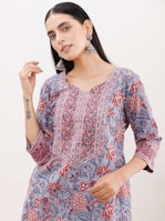A person wearing a floral-patterned kurta with vibrant colors of red, pink, blue, and gray. The outfit features intricate embroidery along the neckline. The individual is accessorized with large, ornate silver earrings and has long, dark hair. The person has a tattoo on their forearm.
