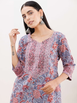 A person wearing a floral-patterned kurta with vibrant colors of red, pink, blue, and gray. The outfit features intricate embroidery along the neckline. The individual is accessorized with large, ornate silver earrings and has long, dark hair. The person has a tattoo on their forearm.