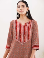 A woman wearing a patterned kurta with a geometric design in shades of red, orange, and white. The kurta features intricate patterns and gold embroidered accents, and she accessorizes with large, dangling silver earrings. Her hair is straight and parted in the middle, and she stands against a plain background.