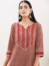 A woman wearing a patterned kurta with a geometric design in shades of red, orange, and white. The kurta features intricate patterns and gold embroidered accents, and she accessorizes with large, dangling silver earrings. Her hair is straight and parted in the middle, and she stands against a plain background.