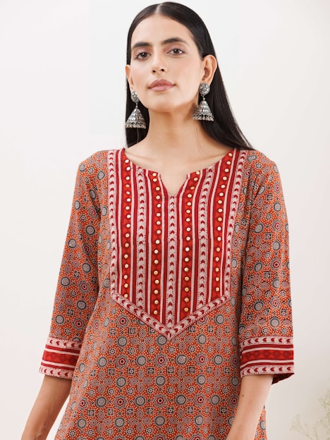 A woman wearing a patterned kurta with a geometric design in shades of red, orange, and white. The kurta features intricate patterns and gold embroidered accents, and she accessorizes with large, dangling silver earrings. Her hair is straight and parted in the middle, and she stands against a plain background.