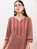 A woman wearing a patterned kurta with a geometric design in shades of red, orange, and white. The kurta features intricate patterns and gold embroidered accents, and she accessorizes with large, dangling silver earrings. Her hair is straight and parted in the middle, and she stands against a plain background.