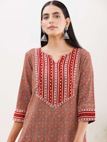 A woman wearing a patterned kurta with a geometric design in shades of red, orange, and white. The kurta features intricate patterns and gold embroidered accents, and she accessorizes with large, dangling silver earrings. Her hair is straight and parted in the middle, and she stands against a plain background.