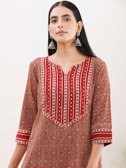 A woman wearing a patterned kurta with a geometric design in shades of red, orange, and white. The kurta features intricate patterns and gold embroidered accents, and she accessorizes with large, dangling silver earrings. Her hair is straight and parted in the middle, and she stands against a plain background.