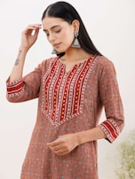 A person wearing a patterned orange and red kurta with intricate geometric designs. The kurta features vertical stripes and decorative elements on the neckline and sleeves. The person is adorned with large, silver jhumka earrings and has a tattoo on their forearm. The background appears to be plain and light-colored.