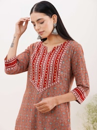 A person wearing a patterned orange and red kurta with intricate geometric designs. The kurta features vertical stripes and decorative elements on the neckline and sleeves. The person is adorned with large, silver jhumka earrings and has a tattoo on their forearm. The background appears to be plain and light-colored.