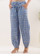 A person is wearing blue patterned pants with elastic cuffs and side pockets. The fabric has a repetitive decorative design featuring dark and light motifs. The individual is also wearing beige high-heeled sandals. The background is a neutral light color, and there is a small plant visible on the side.