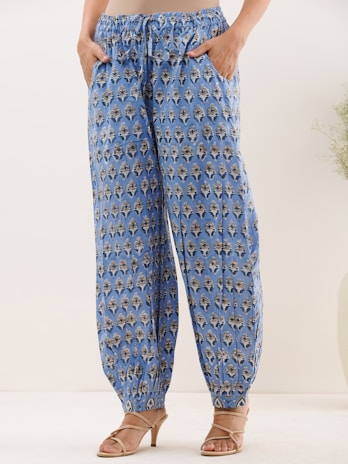 A person is wearing blue patterned pants with elastic cuffs and side pockets. The fabric has a repetitive decorative design featuring dark and light motifs. The individual is also wearing beige high-heeled sandals. The background is a neutral light color, and there is a small plant visible on the side.