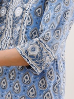 A close-up of a fabric featuring intricate floral and paisley patterns in blue and beige, with a visible portion of a person's arm wearing a garment made of the same fabric.