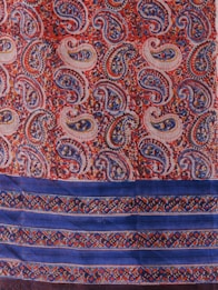 A vibrant textile pattern featuring an array of paisley shapes in rich colors including red, blue, and orange covers the upper portion. The design is intricate with detailed floral motifs inside each paisley. The lower portion of the textile displays horizontal blue stripes embellished with colorful, geometric patterns.