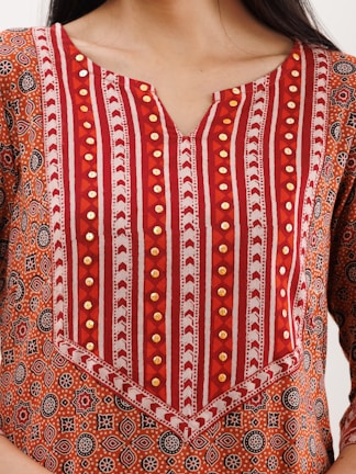 A close-up of a garment with intricate patterns featuring red, orange, and white stripes, along with circular geometric motifs. The fabric is embellished with gold-colored decorative elements.