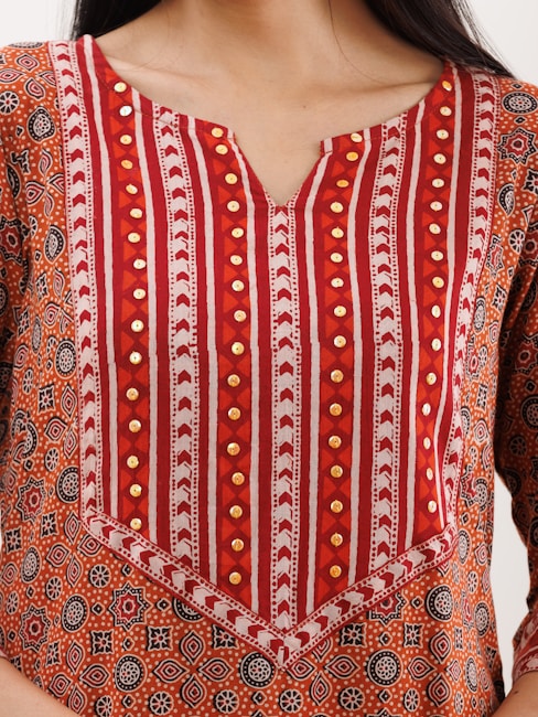 A close-up of a garment with intricate patterns featuring red, orange, and white stripes, along with circular geometric motifs. The fabric is embellished with gold-colored decorative elements.