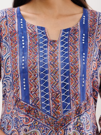 An intricately designed fabric with a mix of paisley and geometric patterns in vibrant colors. The top features vertical blue panels with crisscross white lacing and decorative sequins. The fabric has a colorful combination of red, orange, and white accents.